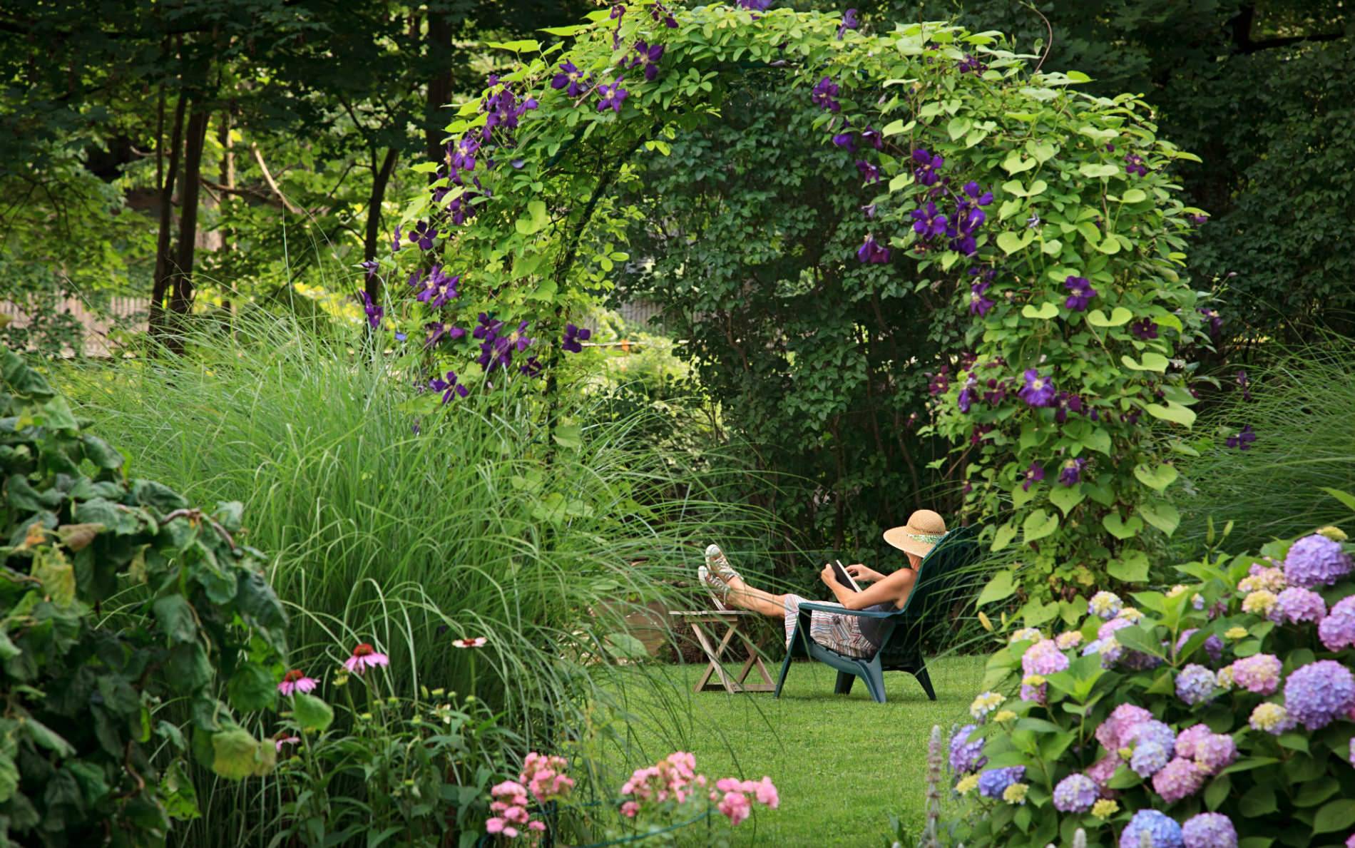Woman relaxing in an Adirondack chair surrounded by green grass and lush green shrubs, flowers and trees