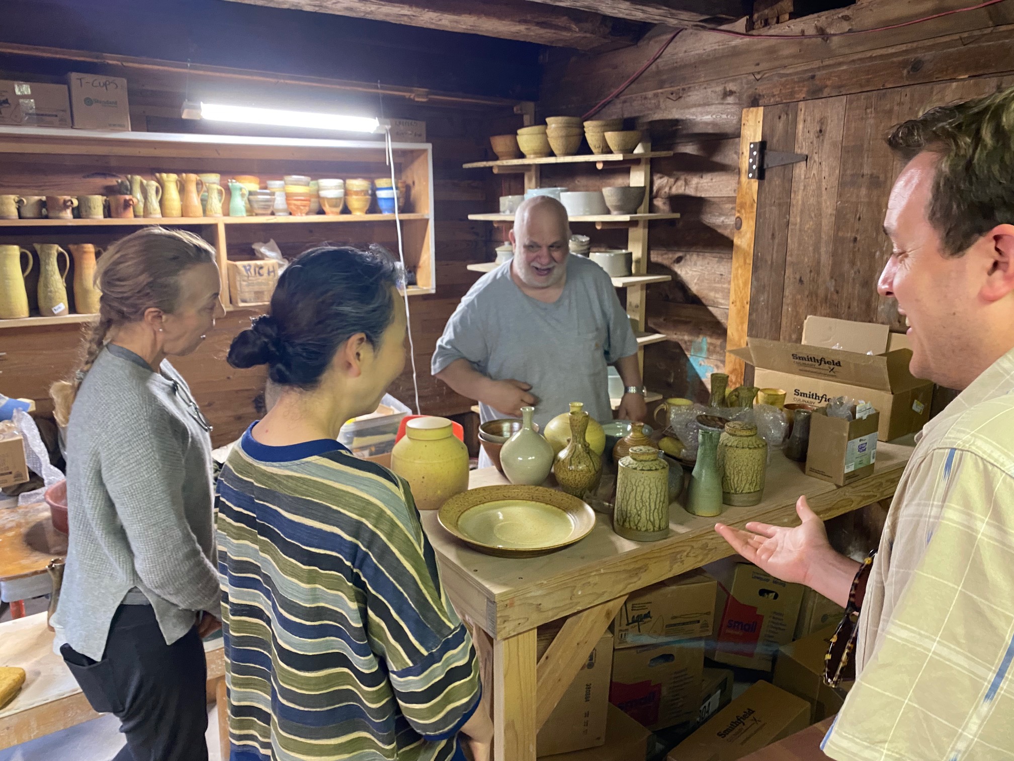 three peopel standing around a table while a man in blue t-shirt talks about the ceramic pieces on the table